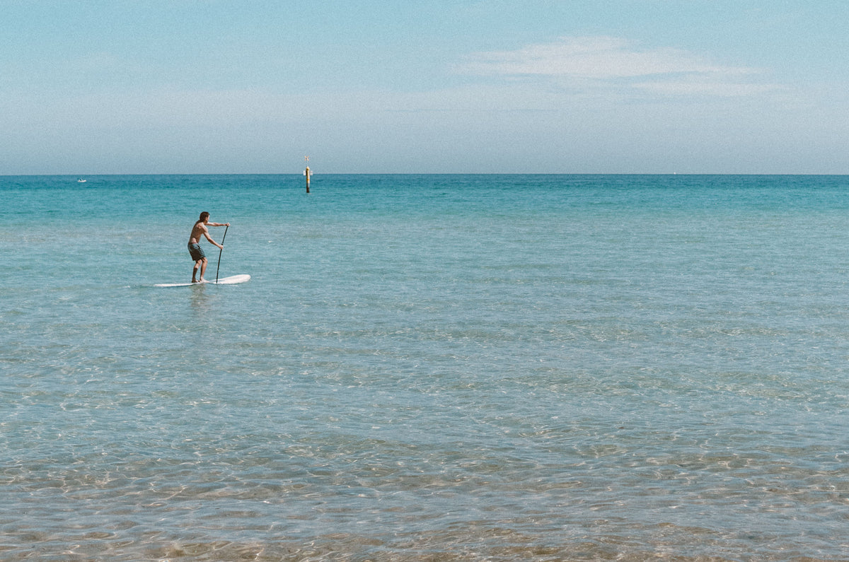 A man enjoying paddle boarding on calm, clear ocean waters under a bright blue sky.